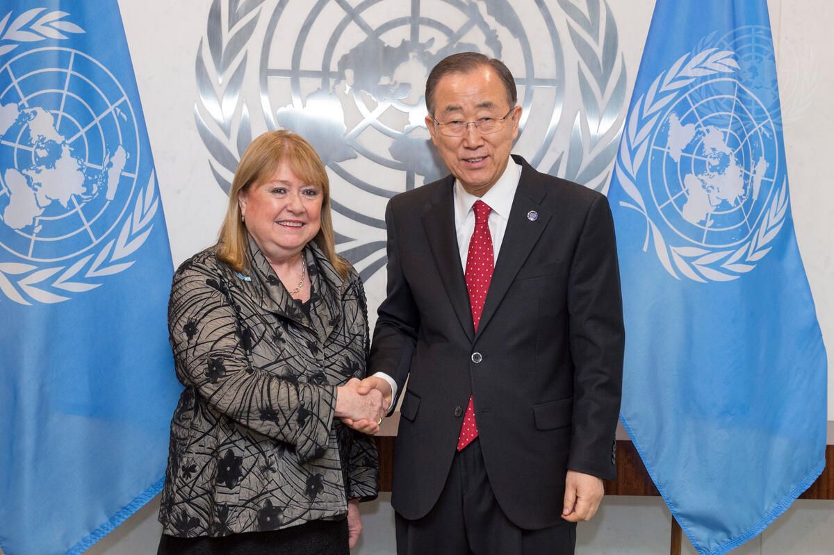 Two people shaking hands in front of UN emblem and between two UN blue flags.