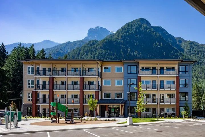 A modern apartment building with a playground and parking lot. The building is surrounded by mountains and trees.