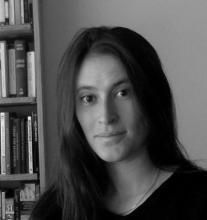 Black and white headshot of Leah Zamore. Person with long dark hair in front of a bookshelf.