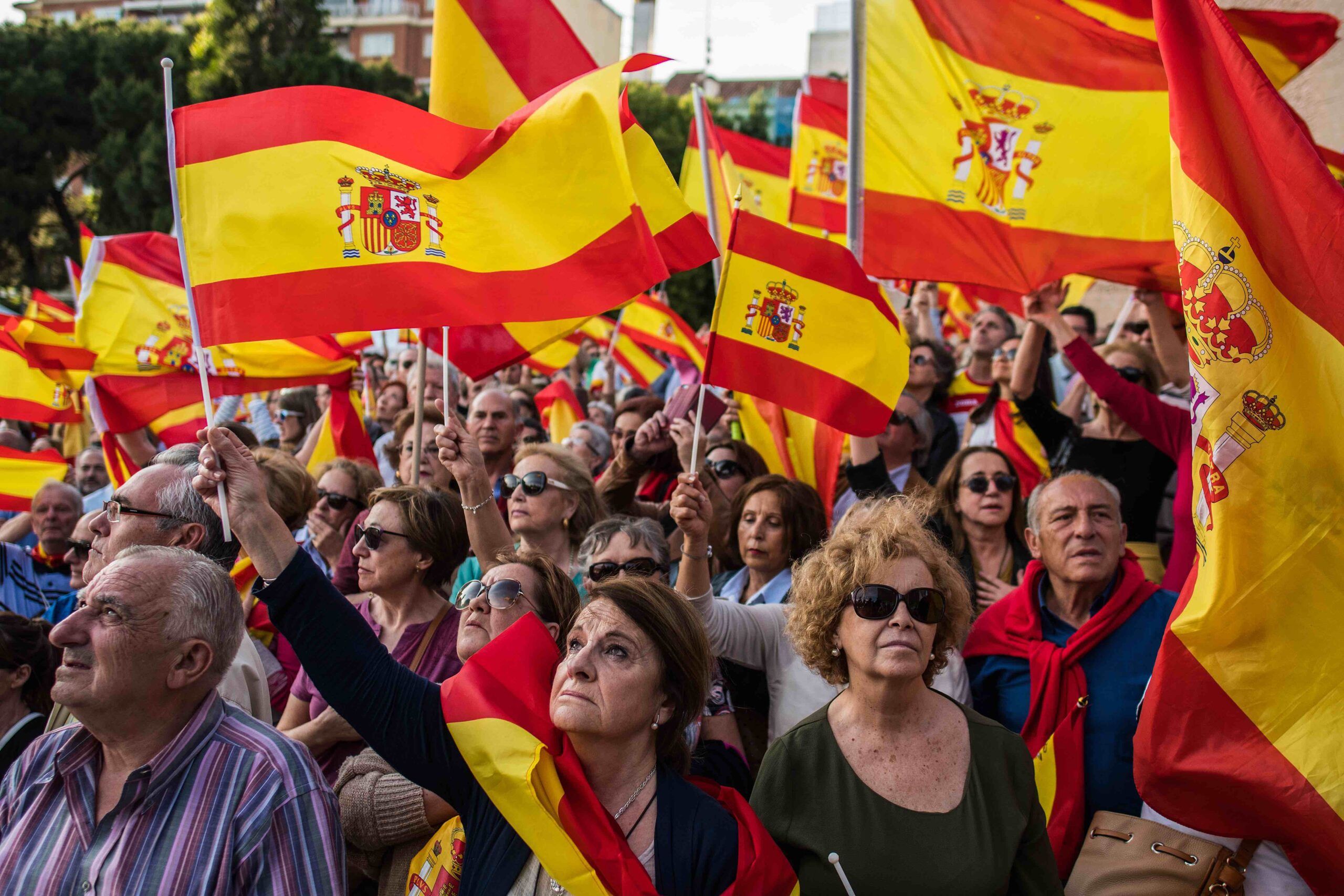 Photo of people waving Spanish flags during a protest demanding general elections and against Pedro Sanchez's Government after motion of censure, in Madrid, Spain. The crowd is looking towards the left, with a few people looking towards the right in the bottom corner of the photo.