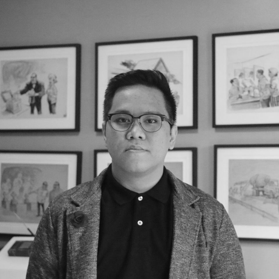 Medium shot of Vino Lucero, a young man wearing glasses, a black polo and a grey cardigan standing in front of a wall with framed pictures