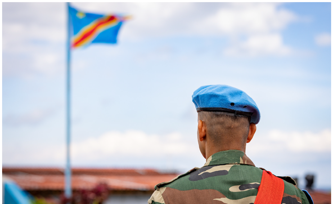 MONUSCO solider dressed in ceremonial uniform looking at a Democratic Republic of Congo flag on a flagpole.