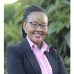 Headshot of a woman with glasses, pink collared shirt, and black blazer smiling in front of a blurred outdoor background.