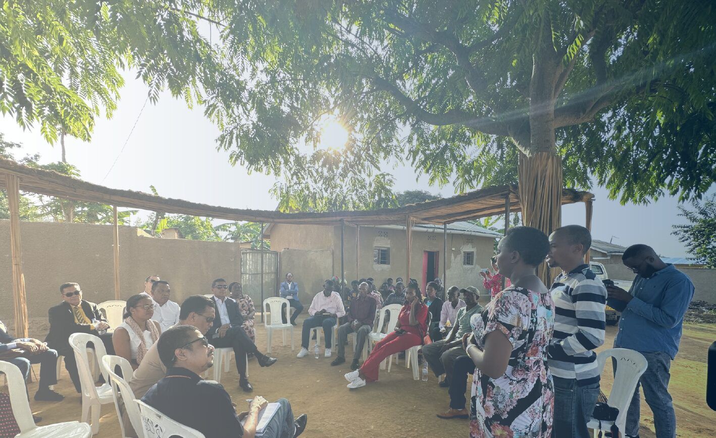 Group of men and women sitting outdoors in white plastic chairs arranged in a circle beneath a rudimentary veranda and a sprawling tree