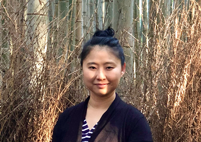 Headshot of an Asian woman standing in a forest, wearing a black and striped top.