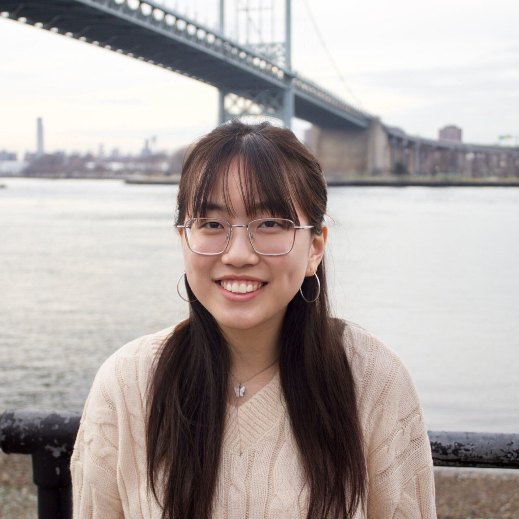 Asian person with glasses and long brown hair standing in front of a riverfront and bridge.