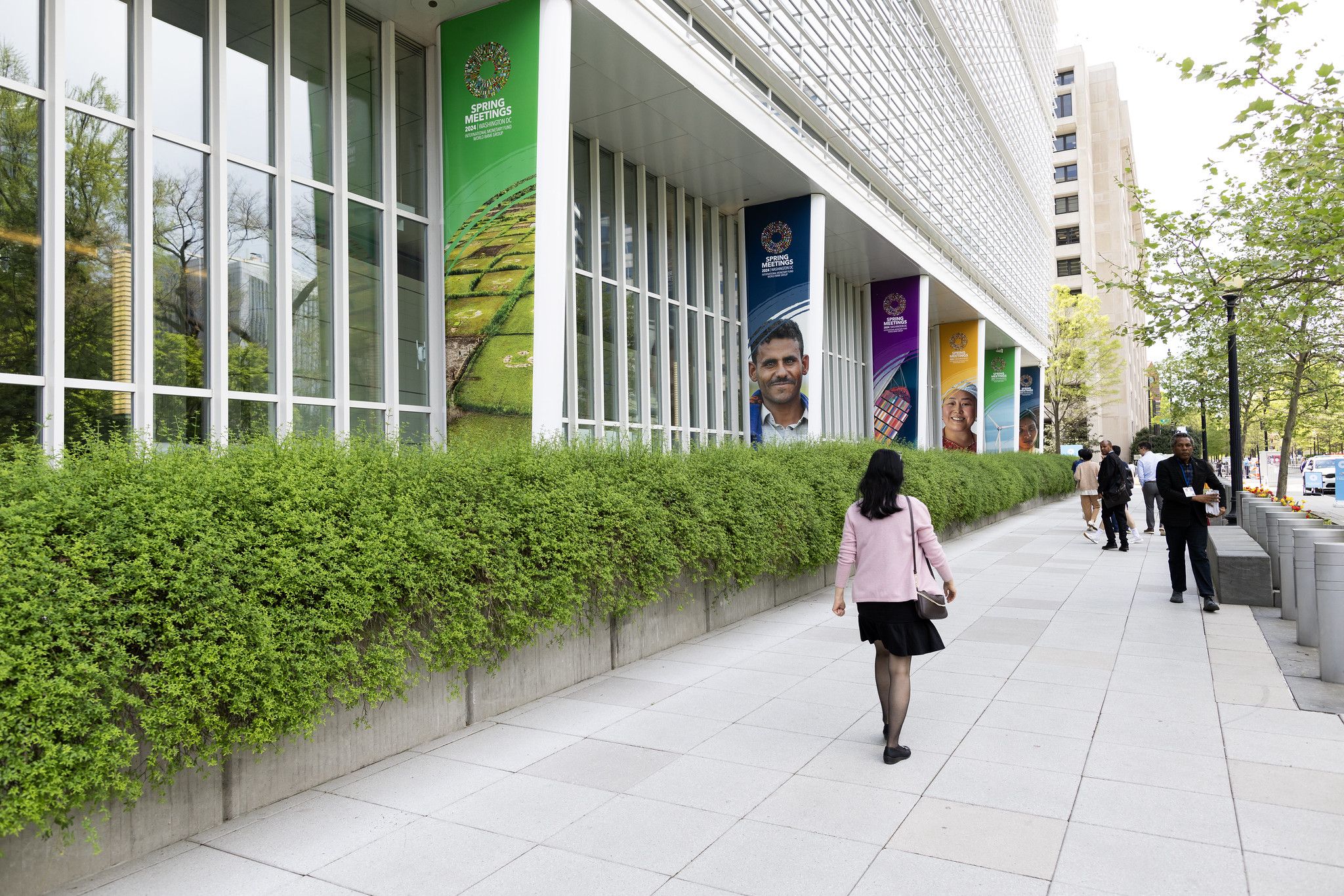 World Bank Group headquarters exterior with Spring Meetings signage and a lush green hedge. People are walking on the sidewalk with their back facing the photographer.