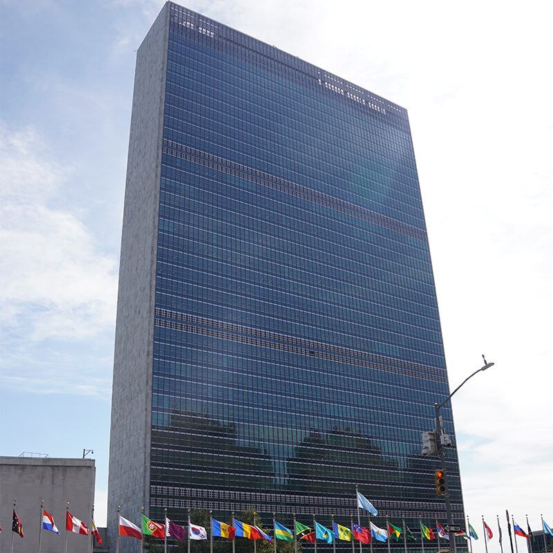Tall UN building against the blue cloud sky. At the base are different flags flying.