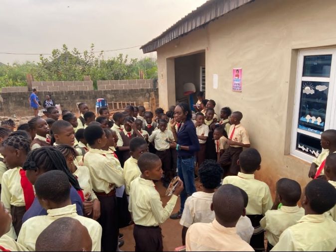 Group of school children looking at the facilitator in front of a schoolhouse.