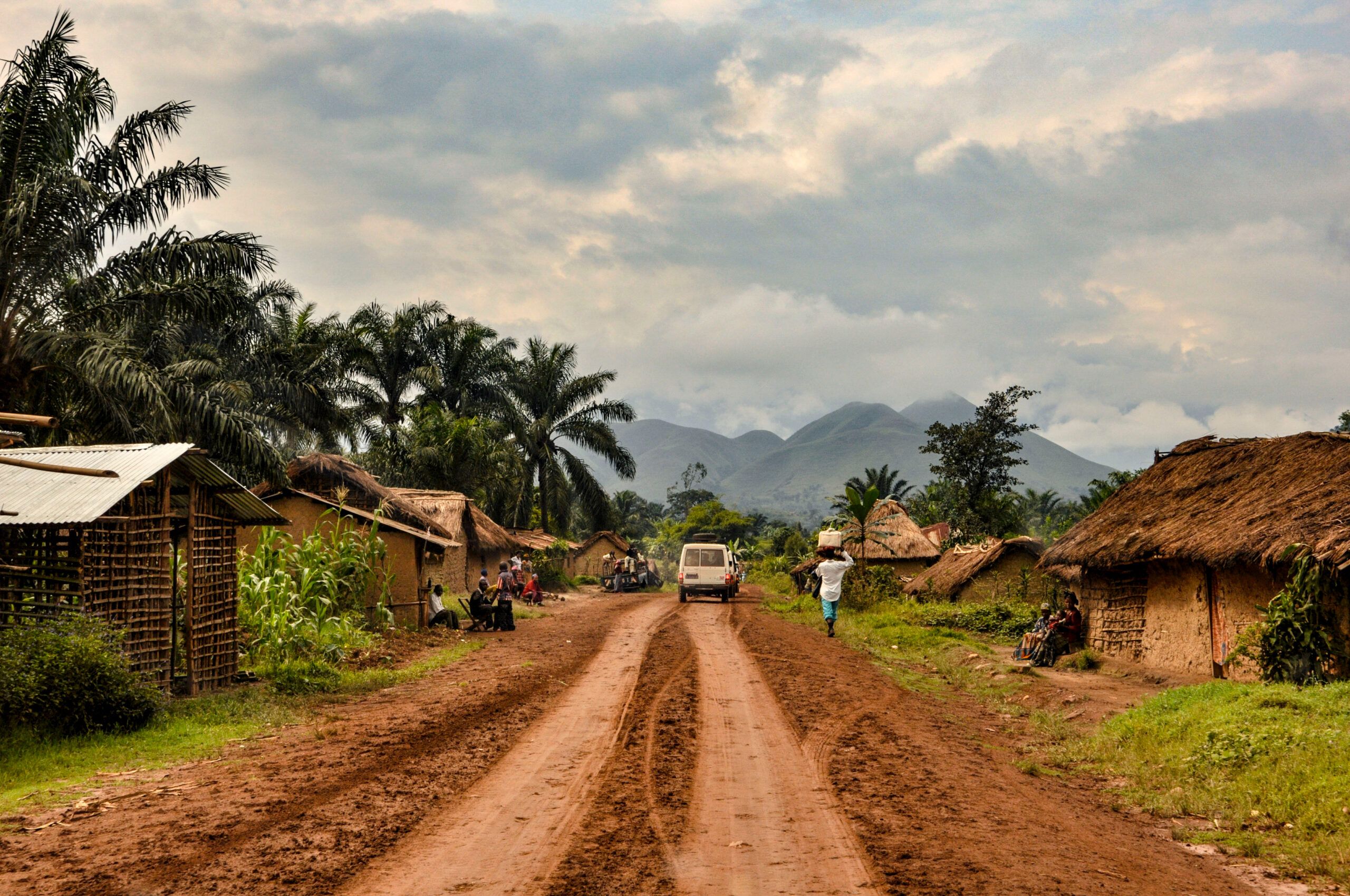 This image depicts a rural village scene with a dirt road running through the center. On either side of the road are thatched huts and simple wooden houses, surrounded by lush greenery and palm trees. A vehicle is traveling down the road, and people are walking or sitting near the homes. In the background, there are misty, rolling mountains under a cloudy sky, adding to the peaceful yet remote atmosphere of the village.