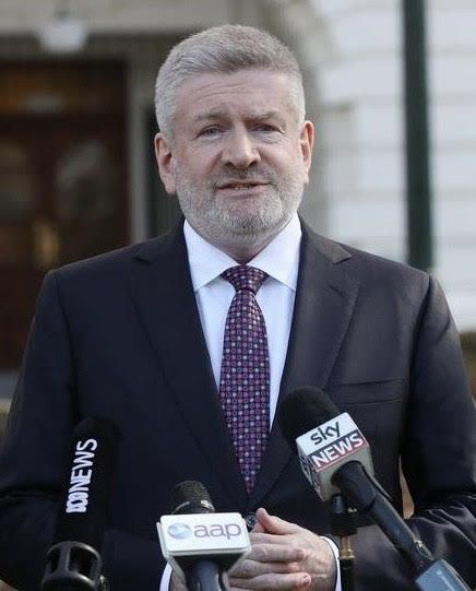 Headshot of a person in a black suit speaking into microphones outside