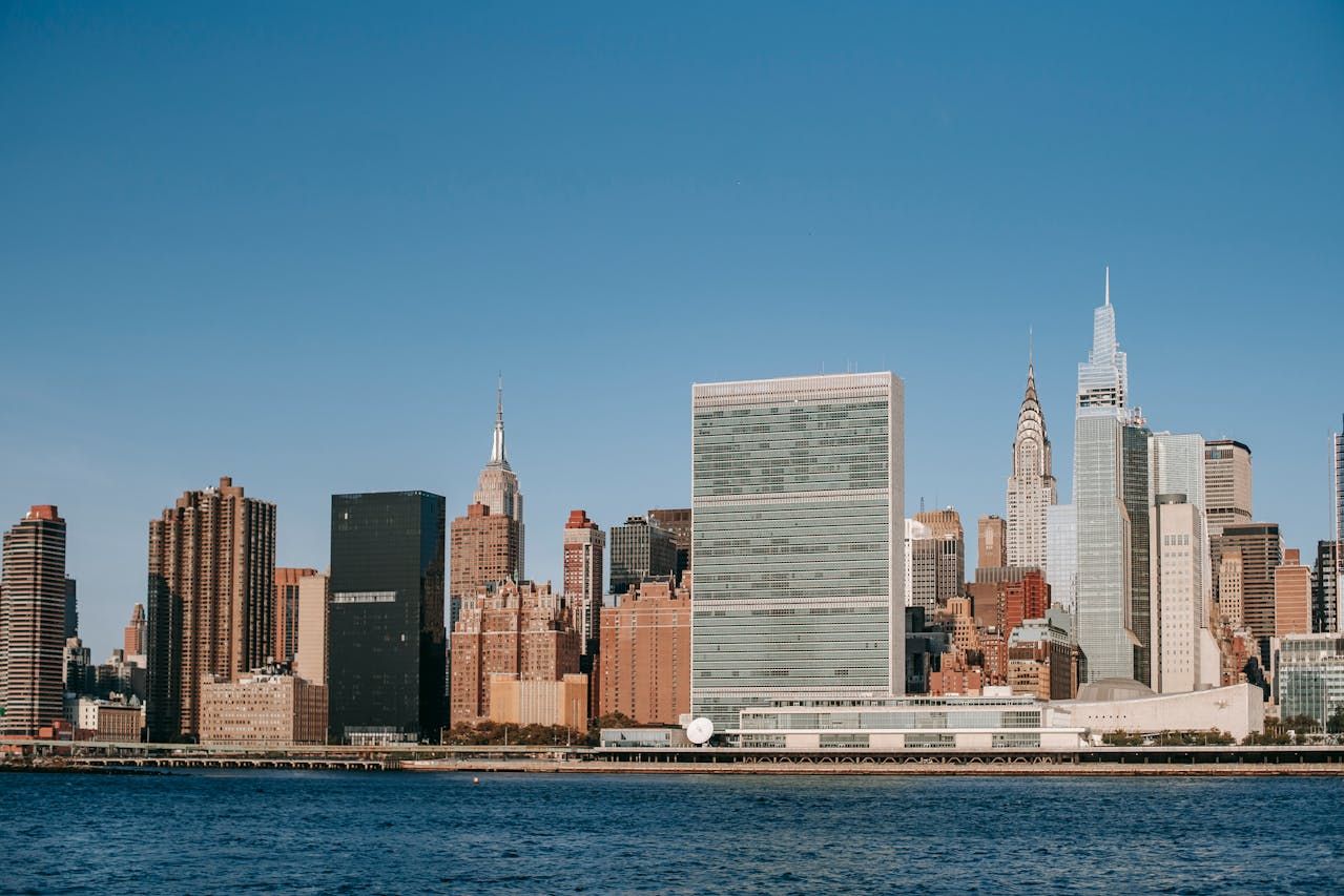 Row of buildings on the waterfront of a river, including prominent New York City skyscrapers such as the Chrysler Building, the Empire State Building, and the United Nations headquarters.