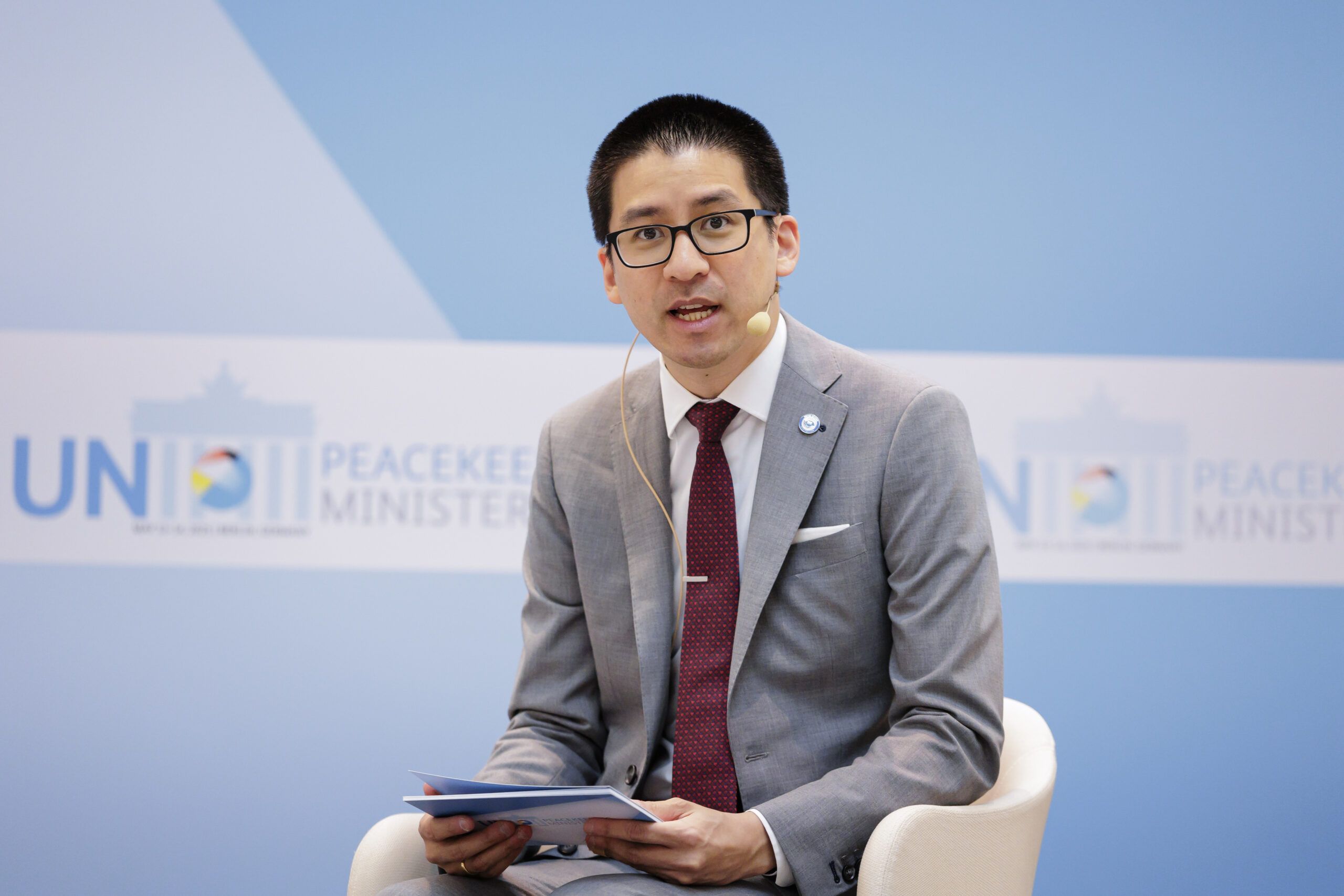 Seated person in a gray suit and red tie and glasses speaking into a mike.
