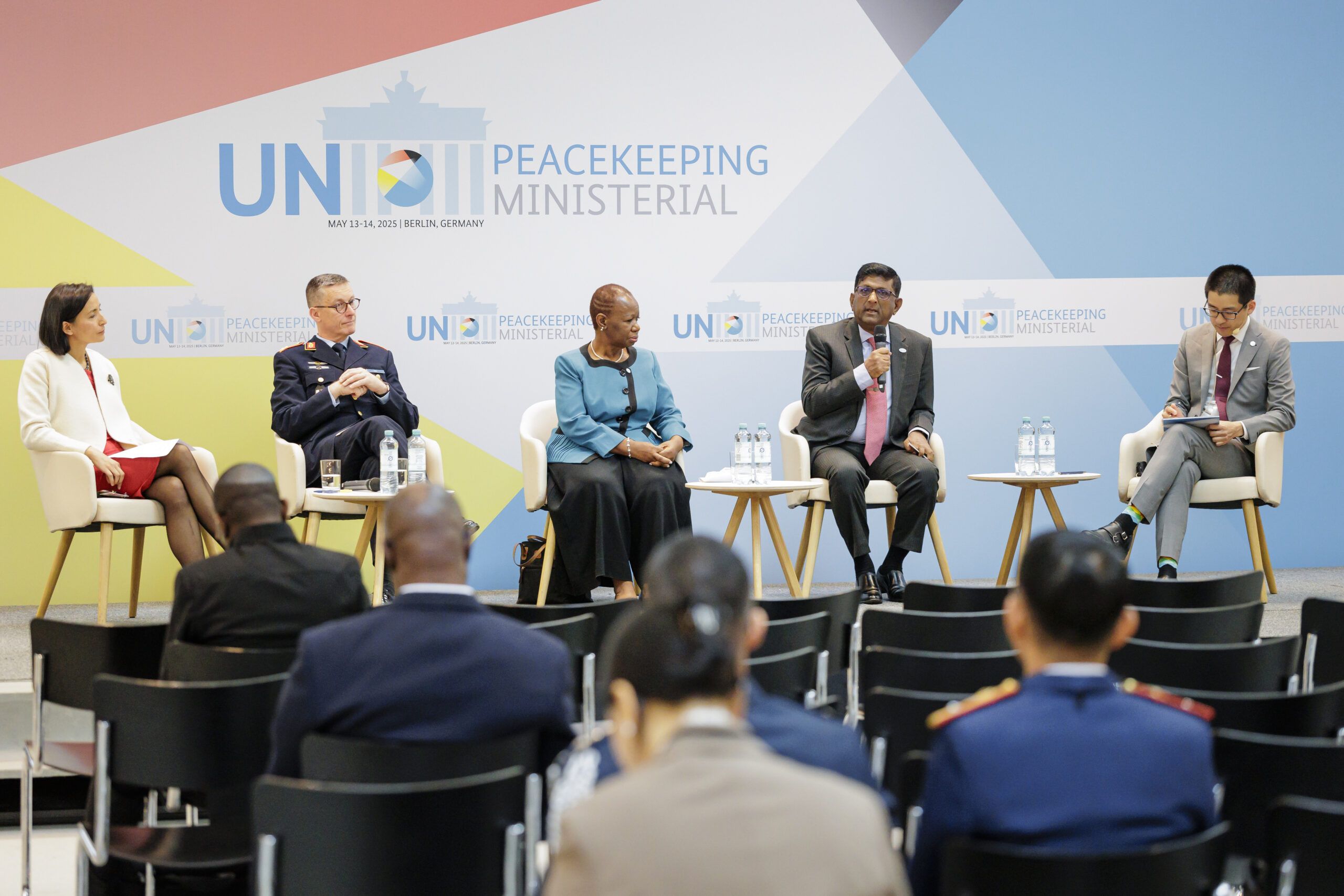 Five individuals seated in front of a colorful stage speaking out to an audience.