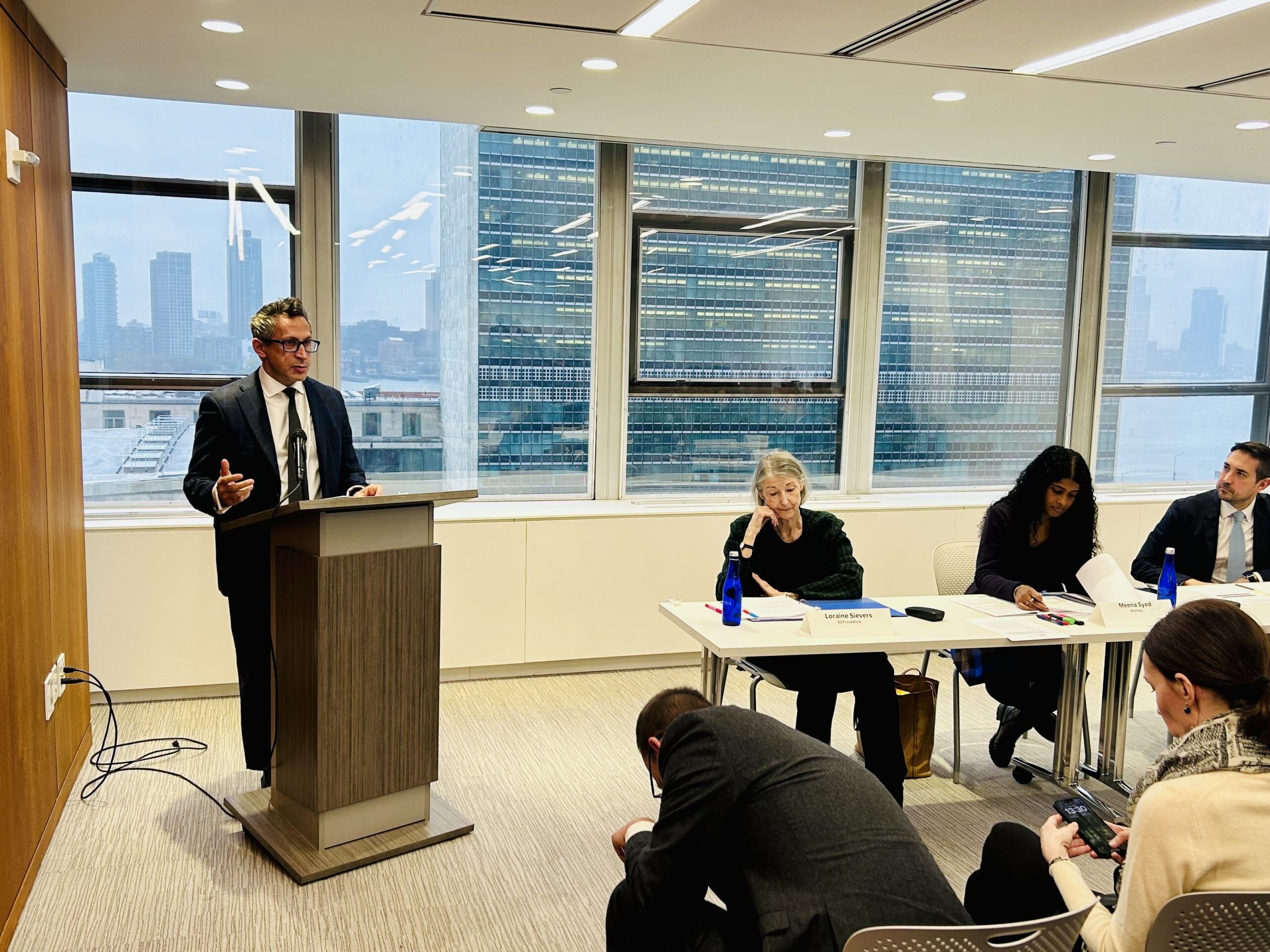 Person standing at a podium speaking in front of windows facing the UNHQ in New York.