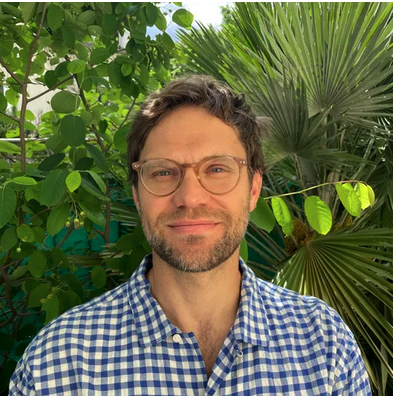 Headshot of a person with a plaid dress shirt, glasses, and brown hair against a leafy green background.