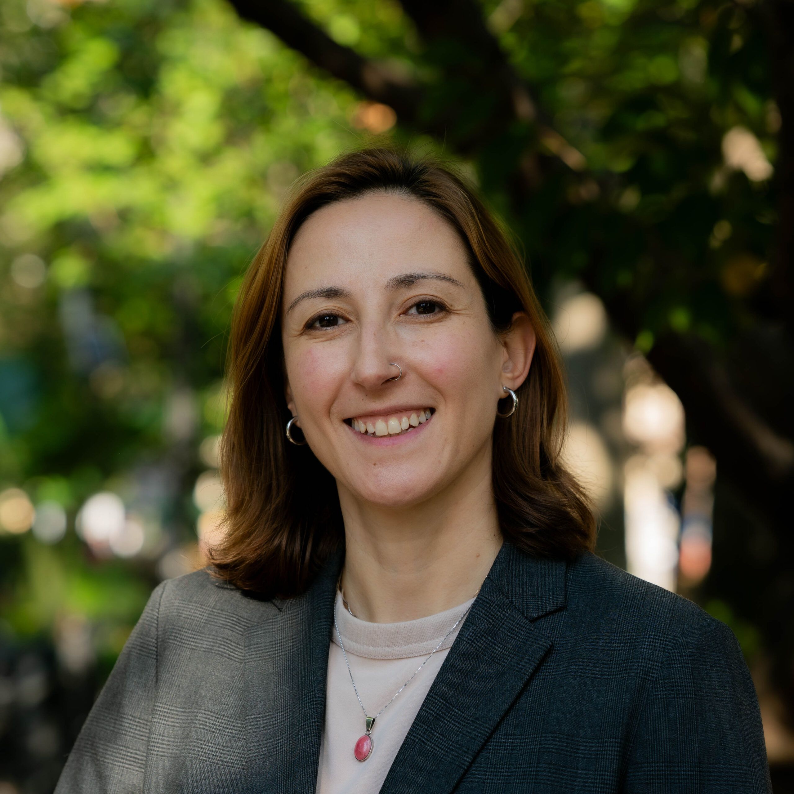 Headshot of a person wearing a gray blazer with short brown hair outdoors. Trees are seen behind her.