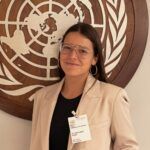 Headshot of a person posing in front of the UN logo. She is wearing glasses and a beige blazer.