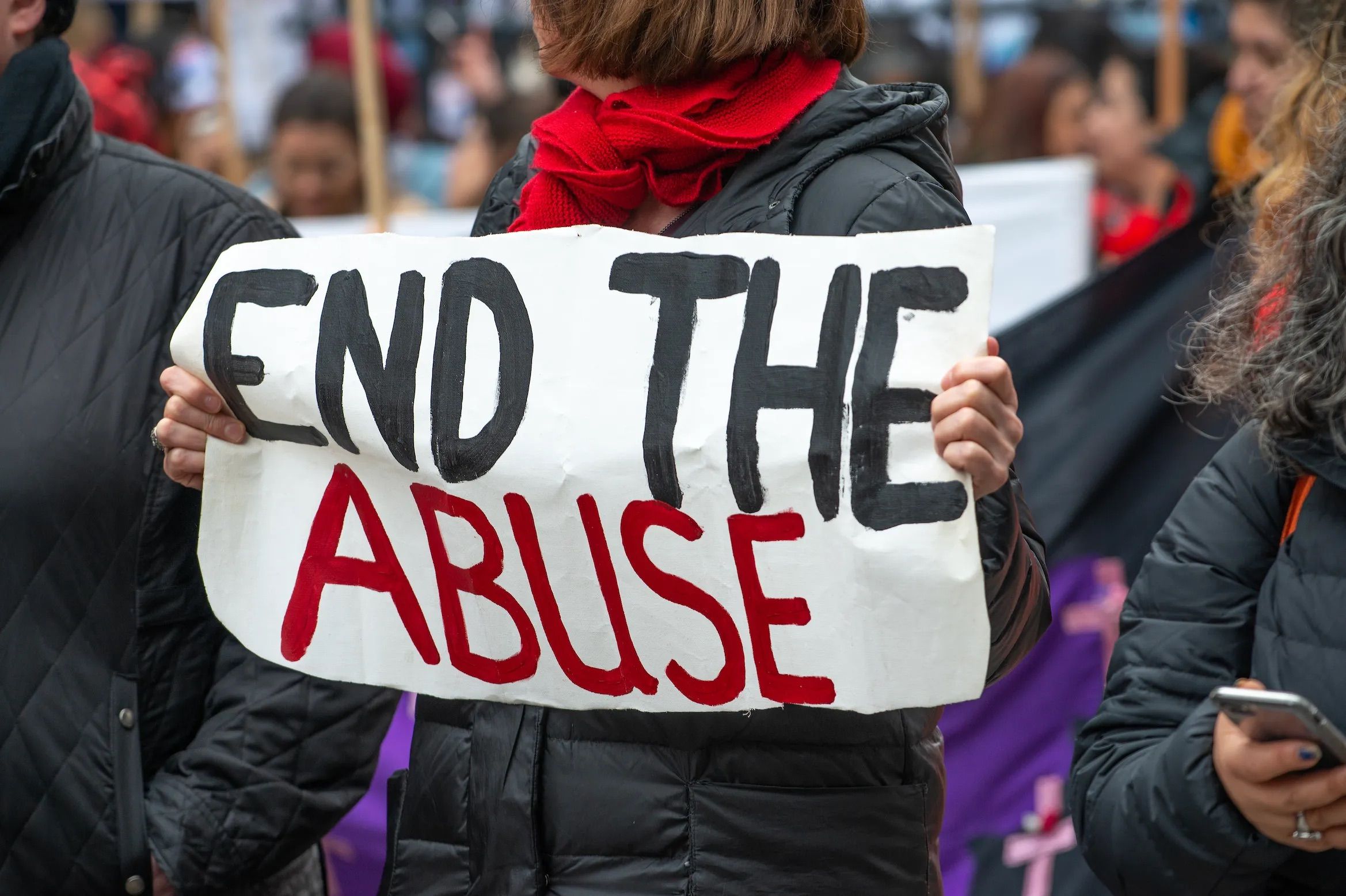 A person wearing a black jacket and a red scarf holds a handmade sign reading 