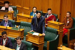 Young Justice Leader Shaneel Lai standing up and speaking at a youth parliamentary meeting. Participants are looking at the speaker.
