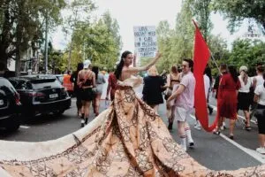 Activists walking down a street at a protest. Focus is on a queer Indigenous youth wearing an elegant traditional dress and holding a sign: "Love doesn't need a cure. End religious conversion therapy."