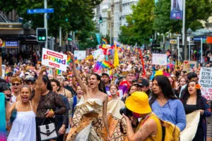 Crowd of protesters filling up a street, holding up Pride flags and protest signs. Activist leaders are standing at the front with their fists raised.