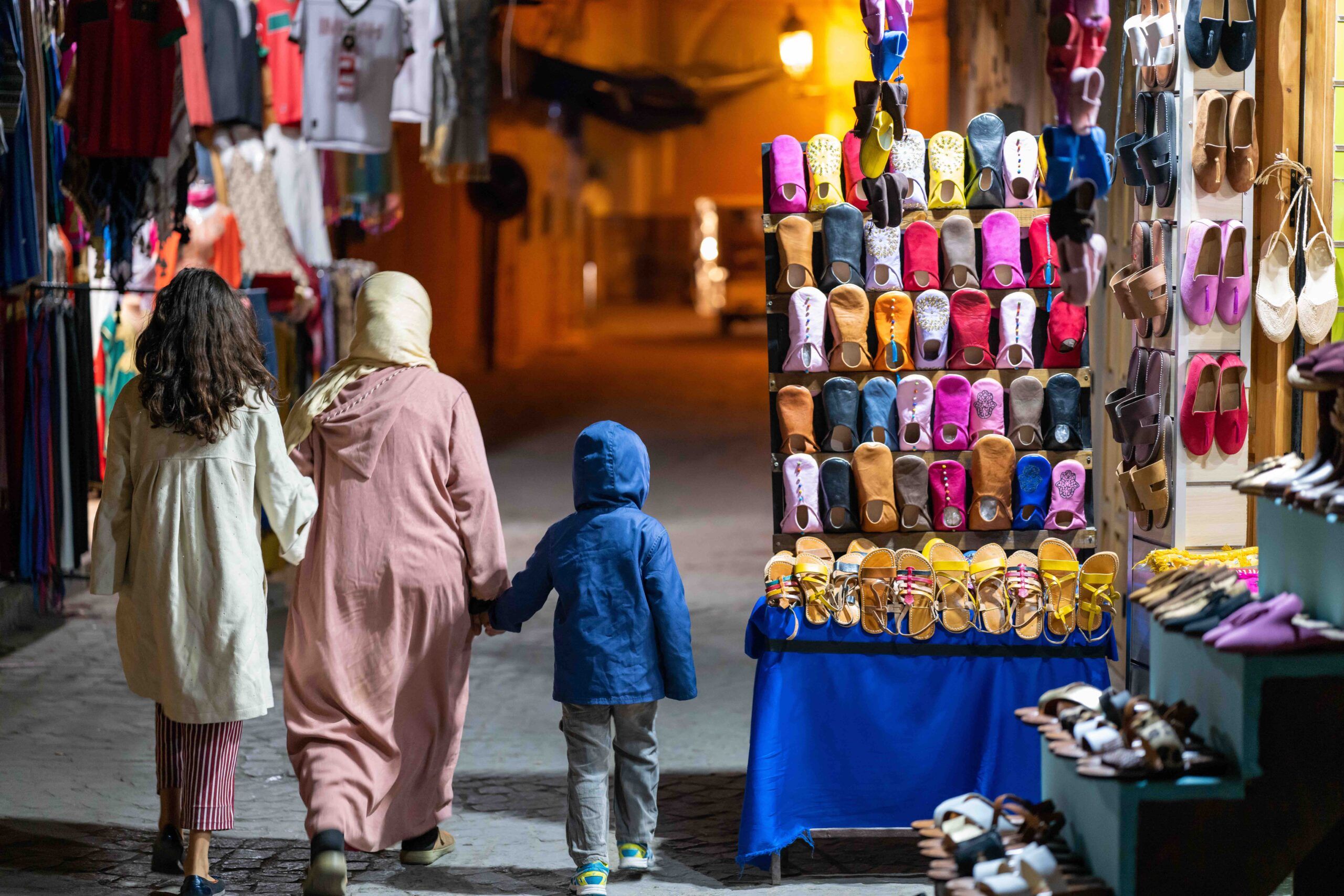Two people and a child walking in a street market in Marrakesh with their backs towards the camera. One of the adults has brown hair, and the other has a headscarf. The child is wearing a blue jacket.