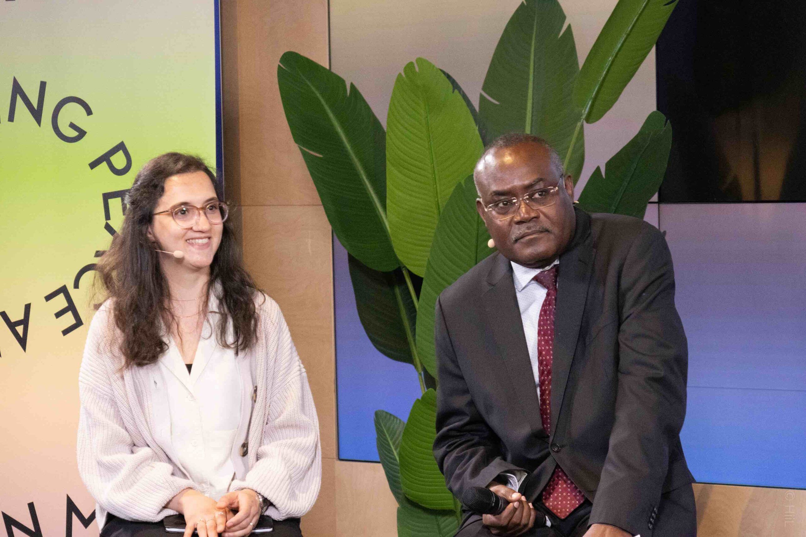 Two people sitting in front of a banner and large plant at the Hague Justice Week 2023.