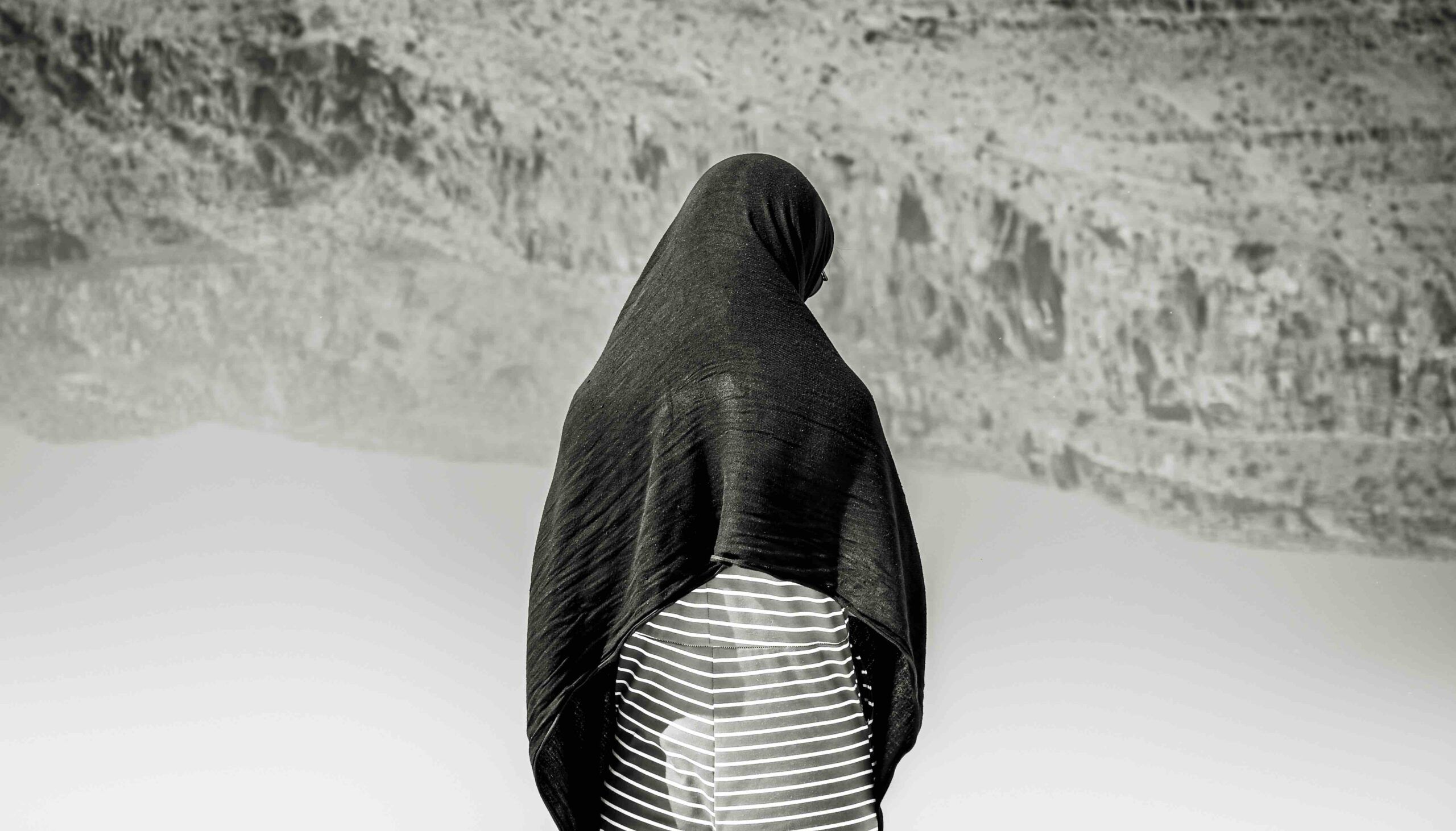 Black and white photo: back view of a woman wearing a hijab standing alone looking at a lake