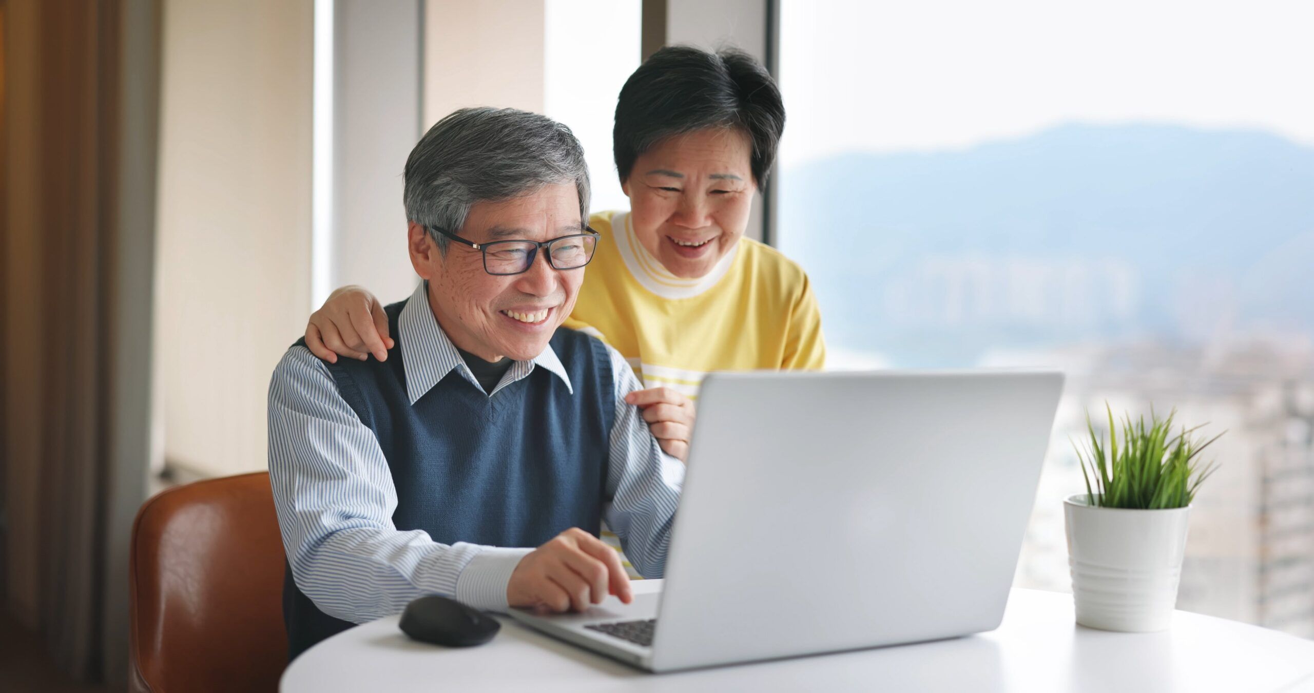 Asian senior couple are smiling and using a laptop near a window, with mountainous landscape in the background.