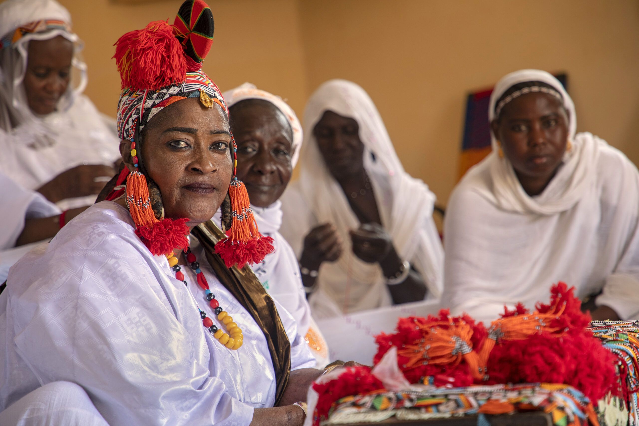 Women gathered in white clothing and headscarves, with the leader facing the camera with a bright red/orange ornamental headress on.