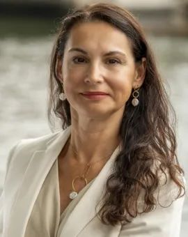 Headshot of a person with long brown hair and earrings wearing a white blazer.