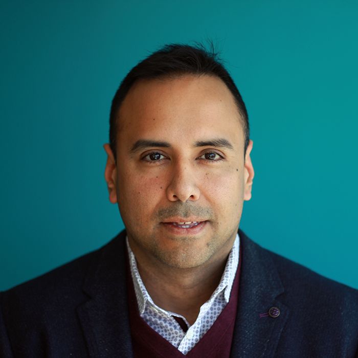 Headshot of a person with short hair wearing a black sweater over a collared shirt on a teal background.