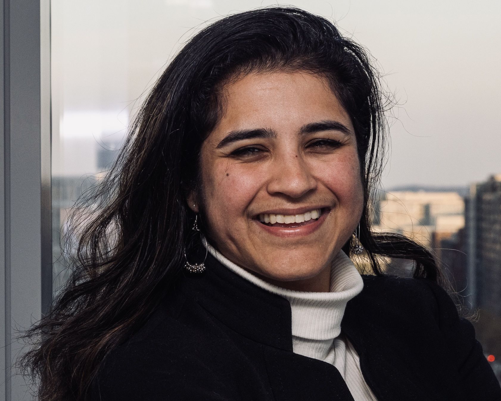 Headshot of a person with dark brown hair wearing a black collared jacket over a white turtleneck.