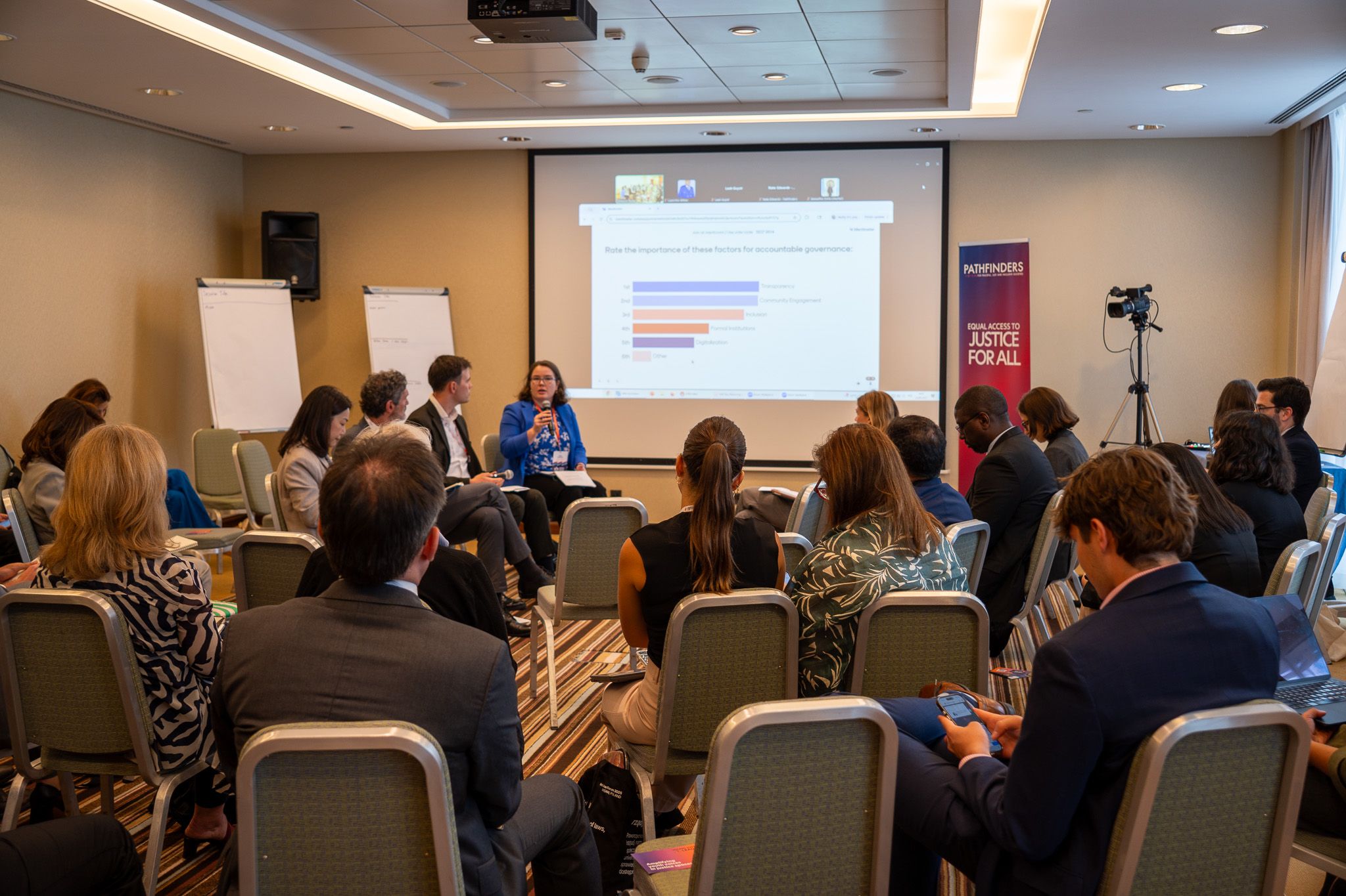 Group of participants listening to a hybrid panel session in a room with a screen at the front.